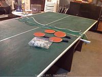 Photo showing green ping pong table surface with the net and 4 paddles, ping pong balls on the table, table worn in several areas.