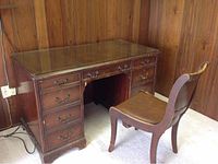 Photo showing antique wood desk and chair set in wood paneled room with carpet underfoot. Desk has glass top over leather surface and shows scratches and wear. Chair has leather seat and back with minor wood scratches.