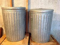 Photo showing two large vintage galvanized metal trash cans with fitted lids, placed side by side on a wooden surface.