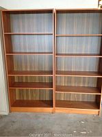 Two tall pressed wood bookshelves side by side against a wall in a room with concrete floor. Each bookshelf has four adjustable shelves and a wood veneer back panel.