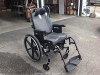 Black tilt-back wheelchair upright showing seat, adjustable headrest, armrests, rear and front wheels, and footrests.