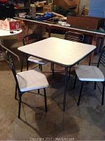Two 34" x 34" white vinyl padded card tables with charcoal metal frames and four matching metal chairs with white vinyl padded seats set up in a garage/workshop space.