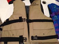 Photo of beige Kent Pro Fisherman kid's life jacket with black straps and pockets alongside blue kid's life jacket with purple straps.