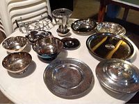 Top-down view of fifteen assorted silver plate and metal serving pieces including bowls, platters, plates, and covered dish arranged on a table.