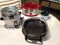 Photo showing Rival Little Dipper crock pot (white with red lid), rice cooker with metal lid, square glass electric skillet with black handles, and bun warmer pouch on round table