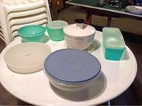 Photo shows six assorted Tupperware pieces including a white lettuce spinner, various round containers, and a rectangular container, all with lids, on a white round table.