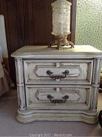 Front view showing one ivory-colored night stand with two drawers, geometric decorative molding, and metal handles, displaying vintage wear and scratches.
