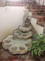 Full view of the three-tier cement water fountain with the figurine on top, showing water flow and size relative to surrounding stairs and wall.