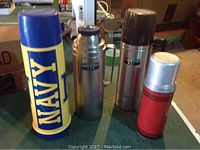 Four thermoses displayed on a table: a large blue one with yellow NAVY logo, two stainless steel ones with black tops, and a smaller red one with a silver top.