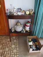 Full view of wooden table with various decorative figurines on top and lower shelf, plus a box with additional items on floor beside it.