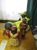 Three cast iron banks and a handmade fabric doll on a wooden table near a window. Visible details include painted features, clothing colors, and doll's cloth texture.