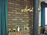 Overall view of brass animal figurines displayed on a brick mantle and a large brass wall plaque of two leaping deer mounted above.