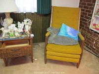 Side view of a wooden side table with decorative drawers and marble inset on top, next to a mustard gold fabric recliner with wooden arms and legs, featuring a blue cushion and gray throw blanket.