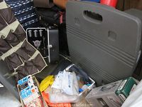 Photo showing a black portable bar case, dark soft sided suitcase, ponchos, and boxed gun cleaning kit on floor