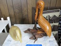 Wide angle image showing both wooden bird sculptures; pelican on driftwood base and smaller duck sculpture beside it on a table.