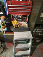 Photo of the Craftsman red tool chest with drawers and some tools inside, small metal ladder placed in front, box of gloves, and boxed Brinkmann outdoor gas cooker visible behind ladder and below tool chest.