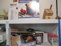 Shelf with Kitchenworks thermal carafe box, Kitchenworks 6-quart electric kettle in box, wooden knife block, Rival electric can opener, Turkey Cannon roasting device, and two Oster ice crushers.