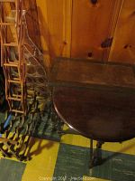 Shows two wooden side tables, one with a rounded top, the other rectangular, placed against a wood-paneled wall with a foldable wood wine rack and acrylic wine rack beside them.