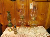 Four oil lamps of varying sizes and glass styles with chimneys, displayed on lace cloth in front of wood paneling, along with a tall electric lamp with fabric shade