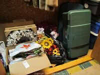 Photo showing white rose wreath, sunflower and other faux flower arrangements partially in boxes and plastic bags next to green plastic storage container