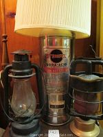 Three lamps side by side: left black metal oil lantern, center extinguisher-based electric lamp with shade, right brass-bottom caged lantern