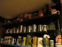 Photo showing multiple shelves of assorted beer steins of various shapes, sizes, and materials including ceramic, metal, and pewter steins.