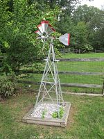 Full view of the metal yard windmill with red and silver painted blades and tail, standing in a yard with fence and greenery in background.