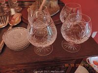 Four cut crystal stem glasses and stack of six round glass coasters on wood table with partial silverware visible