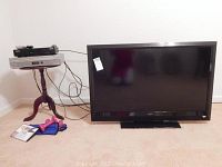 Photo showing the 42 inch Vizio VL420M flat screen TV next to the Zenith DVD-VCR combo player on a small round table, with some cables and small dumbbells on the floor.