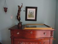 Side view of bronze Athena statue, one framed art print of building in black frame on wall, wooden dresser surface with large gold-trimmed empty frame laying flat.