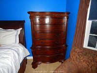 Front view of tall wood dresser next to bed and chair, showing 6 drawers with metal handles and dark polished finish.