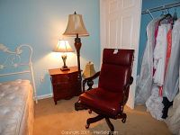 Photo showing two table lamps (one on nightstand with beige tapered shade, one on floor with bell-shaped shade) and a brown leather desk chair with wheels next to bed and nightstand.
