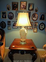 Crystal base table lamp on wooden side table with cream-colored fabric shade against a wall with framed photos.