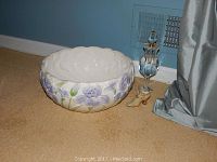 Large white bowl with purple floral design next to crystal perfume bottle and miniature shoe on carpeted floor near curtain.