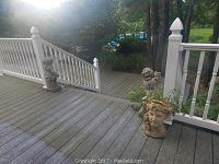 View of three outdoor classical statues on a wooden deck with railing. The bust planter is in front with ivy, and the cherubs are positioned near the railing posts.