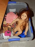 Two American Girl dolls visible in plastic bin, one with red hair nude, and doll clothes including a striped dress.