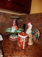 Tiered ceramic appetizer platter with figure on top, Bassano rooster figurine, and floral jar displayed together on countertop.
