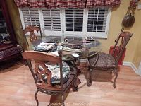 Glass top round dining table with four chairs, two with floral upholstery and two with brown upholstery, positioned on a wood floor next to a window with white blinds and checked curtains.