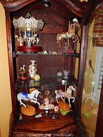 Glass-fronted wooden display cabinet containing carousel horses, decorative china pieces including a floral vase and ornamental dishes on shelves.