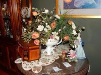 The lot arranged on a glass table showing a porcelain flower figurine, cloisonne bell, Lladro Nao-style figurine, and a wooden box on a beaded lace doily.