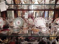 Wide photo of shelf with varied china tea cups, saucers, and glassware inside glass cabinet