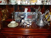 Photo of porcelain decor, crystal bell, tea cup and saucer, Arthur Court pewter tray, and silver-toned pitcher in cabinet