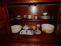 Wide view of china pieces inside a wooden cabinet including bowls, teapot, cups, and plates.