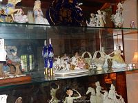 Cabinet shelf showing porcelain figurines, cobalt blue glass bottles, egg baskets and carriage figurine