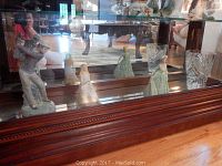 Porcelain figurines and crystal tumblers displayed in a glass cabinet with wooden base, showing four porcelain figurines including a golfer and three female figures, paired with two clear cut crystal tumblers.