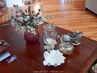 View of various decorative objects on a wooden table including red glass vase with white and pink artificial flowers, clear cut crystal jar with lid, white porcelain shell dishes, and cloisonne butterfly figurine.