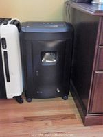 Black Executive Machines paper shredder positioned next to a suitcase and wooden desk, showing front with transparent window and four caster wheels.