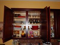 Photo showing open kitchen cabinet with shelves filled with various dishes including red and white ceramic plates, floral and decorated mugs, glass wine glasses, amber goblets, and bowls.