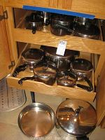 Photo showing multiple stainless steel pots and pans with black handles and lids arranged in wooden drawers and on floor.