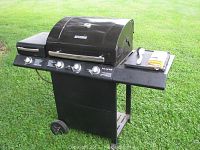 Full view of black Kenmore gas grill on wheels, showing main cooking area with lid, side burner unit on left, and side shelf on right with grilling tools and propane tank connected.